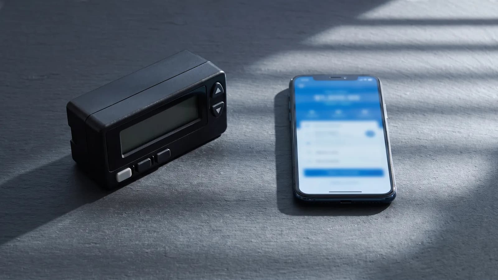 Un compteur horokilométrique de taxi et un smartphone posés côte à côte sur une surface en ardoise, éclairés de la même façon, illustrant les deux modes de tarification.